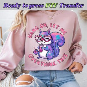 May include: Pink sweatshirt featuring a cartoon squirrel wearing heart-shaped sunglasses and holding a pink drink. The text "Hang on, let me overthink this" is printed around the squirrel.