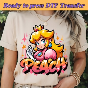 May include: A beige t-shirt featuring a cartoon image of Princess Peach from the Super Mario Bros. video game series. The image is in a graffiti style with the word "Peach" written in pink and yellow letters.