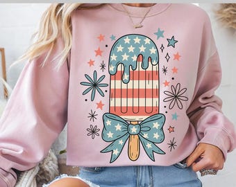 May include: A light pink sweatshirt featuring a patriotic popsicle design with red, white, and blue stars and stripes. The design includes a blue bow and decorative stars and flowers. The text "Ready to press DTF Transfer" is at the top.