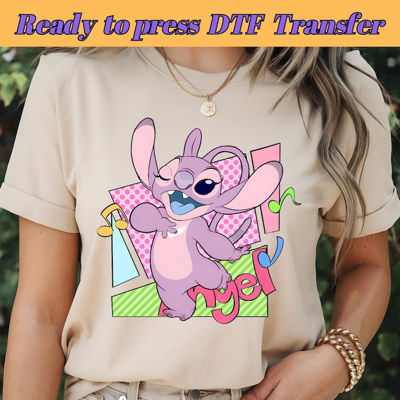 May include: A beige t-shirt with a pink, purple, and blue cartoon character with the word "Angel" printed on the shirt.
