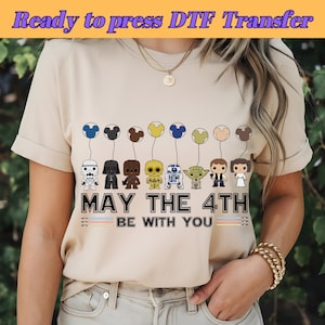 May include: A beige t-shirt with a design featuring Star Wars characters holding balloons shaped like Mickey Mouse heads. The text "May the 4th Be With You" is printed below the characters.