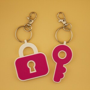 以下が含まれることがあります： シルバー色の留め金とキーリングが付いた2つのキーホルダー。1つはピンクと白の南京錠の形、もう1つはピンクの鍵の形です。背景は無地の明るいオレンジ色です。