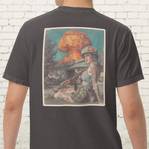 May include: Dark gray t-shirt featuring a vintage-style graphic. The graphic depicts a woman in military attire with tattoos, holding a rifle, and a mushroom cloud in the background. The image has a retro, pin-up aesthetic.