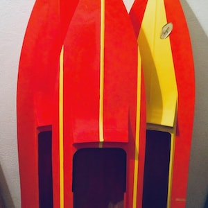 Puede incluir: Tres estanterías de madera con forma de barco. Las estanterías están pintadas de rojo con rayas amarillas en el centro. Una estantería es amarilla y roja. Las estanterías tienen una abertura rectangular. Una pequeña pegatina ovalada está en la estantería amarilla.