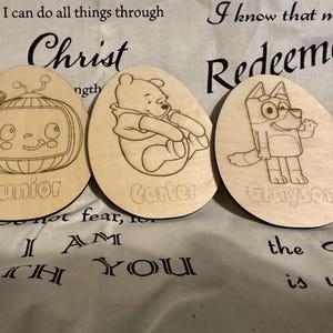 Könnte beinhalten: Drei hölzerne eiförmige Ornamente mit Comicfiguren und Namen. Das erste Ornament zeigt eine Comicfigur aus der Show "Junior" mit dem Namen "Junior" darunter. Das zweite Ornament zeigt Winnie Puuh mit dem Namen "Carter" darunter. Das dritte Ornament zeigt eine Comicfigur aus der Show "Bluey" mit dem Namen "Grayson" darunter.