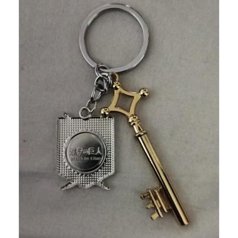 May include: A silver keychain with a gold-colored key and a silver emblem. The key has a diamond-shaped top and a detailed handle. The emblem has Japanese text and a circular design. The keychain ring is silver.
