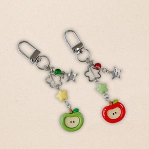 Peut inclure: Deux porte-clés avec des attaches argentées. Chaque porte-clés présente un design de breloques avec une pomme verte ou rouge, des étoiles et des perles en forme de fleur. Les breloques en forme de pomme ont un visage blanc. Les porte-clés sont sur un fond beige clair.