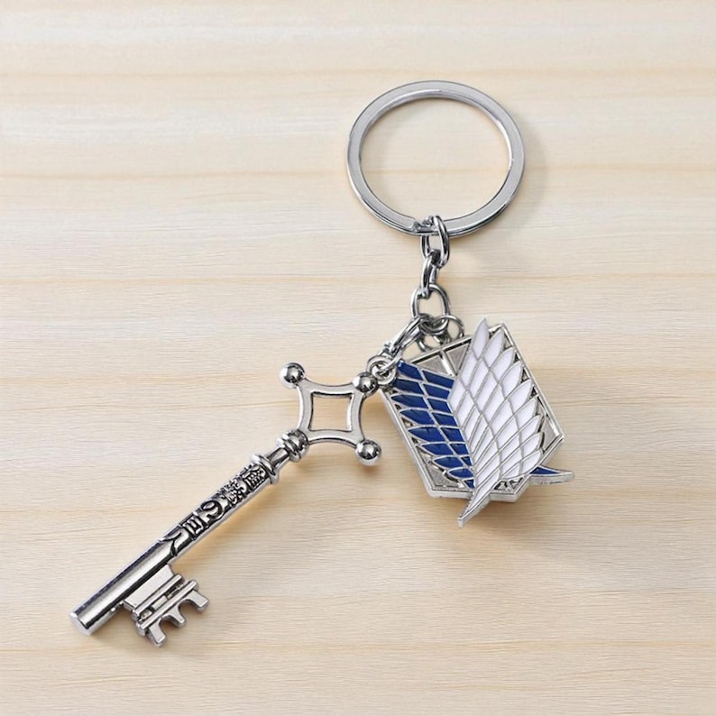 May include: Silver-colored keychain featuring a key and a winged emblem. The key has a detailed design with Japanese text. The emblem is white with blue accents. The keychain is attached to a silver ring.