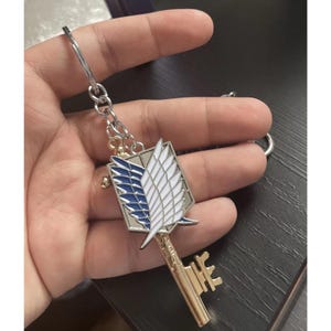 May include: A silver-colored keychain featuring a gold-colored key design. The key has a rectangular emblem with blue and white wings. The keychain is attached to a silver ring and chain.