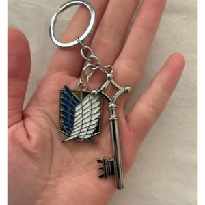 May include: A silver-colored keychain featuring two charms: a key and a shield emblem with blue and white wings. The keychain is attached to a silver ring. The key has a detailed design, and the shield has a gold border.