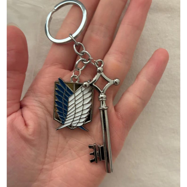 May include: A silver-colored keychain featuring two charms: a key and a shield emblem with blue and white wings. The keychain is attached to a silver ring. The key has a detailed design, and the shield has a gold border.