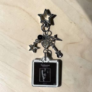 May include: A silver keychain with multiple charms, including stars, a heart, and a guitar. The keychain features a clear, square pendant with a black and white image and the text "The Weeknd Trilogy".
