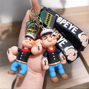 May include: Popeye the Sailor Man keyrings. Each keyring features a Popeye figure with a corncob pipe, sailor hat, and anchor tattoo. The keyrings have black and white Popeye wrist straps and gold-coloured key rings.