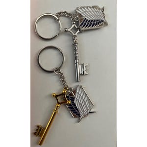 May include: Two keychains with silver and gold keys and a wing emblem. The silver key has a diamond-shaped top and a keyhole design. The gold key has a square top and a keyhole design. Both keychains have a wing emblem with white and blue details.