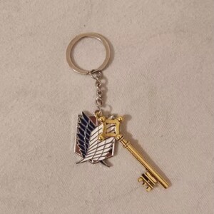 May include: A keychain featuring a gold-colored key and a shield emblem with blue, white, and red details. The keychain has a silver ring and chain. The design is likely inspired by a popular anime or manga series.