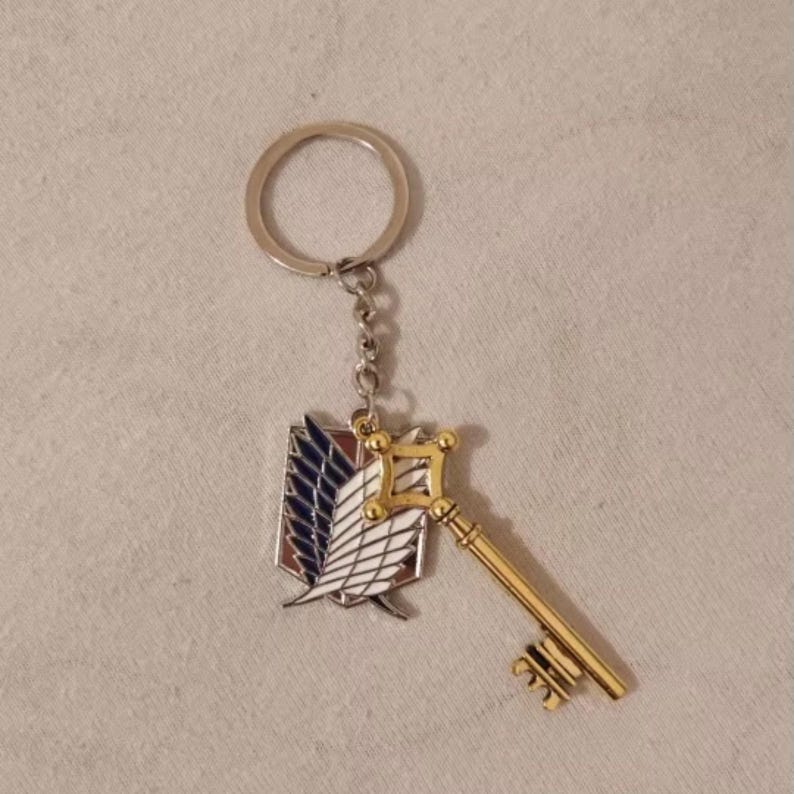 May include: A keychain featuring a gold-colored key and a shield emblem with blue, white, and red details. The keychain has a silver ring and chain. The design is likely inspired by a popular anime or manga series.