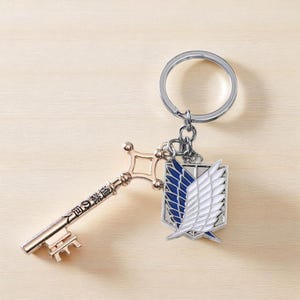 May include: A keychain featuring a gold-colored key with Japanese text and a silver-colored ring. The keychain also includes a pendant with a blue and white wing design.