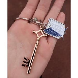 May include: A gold-colored key and a white and blue emblem keychain. The key has a decorative handle and a detailed key bit. The emblem features a wing design. The keychain is attached to a silver ring.