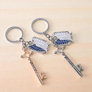 May include: Two keychains featuring a key and a winged emblem design. The keys are metallic, one gold and one silver, with Japanese text. The emblem is white, blue, and silver. The keychains are attached to silver key rings.