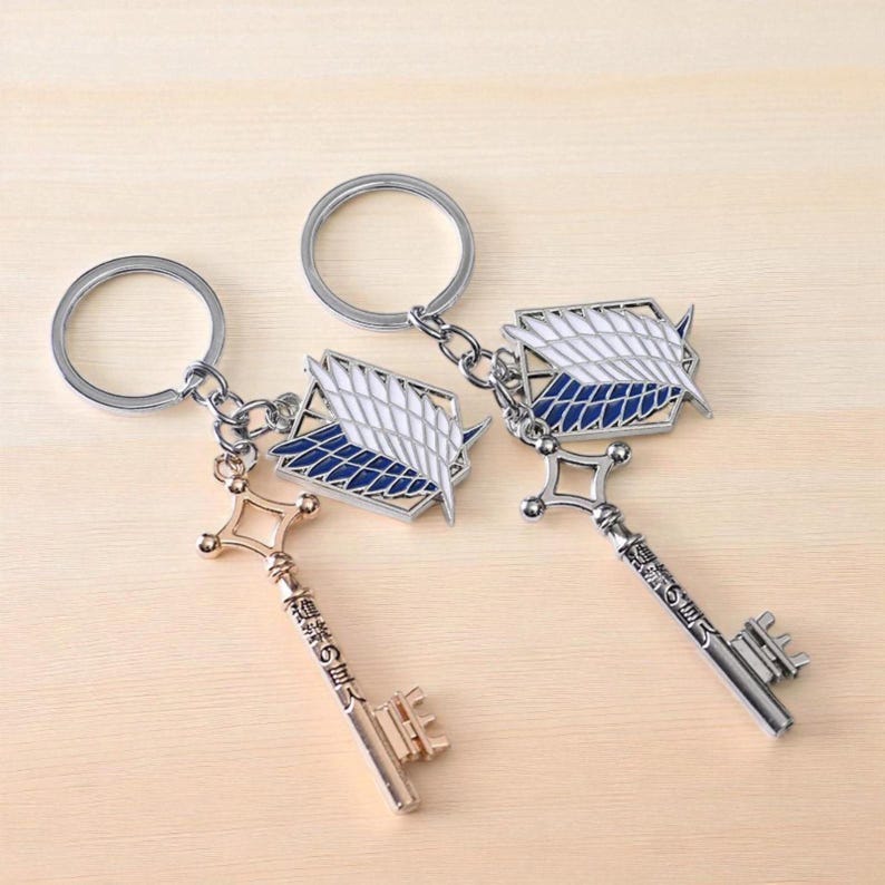May include: Two keychains featuring a key and a winged emblem design. The keys are metallic, one gold and one silver, with Japanese text. The emblem is white, blue, and silver. The keychains are attached to silver key rings.