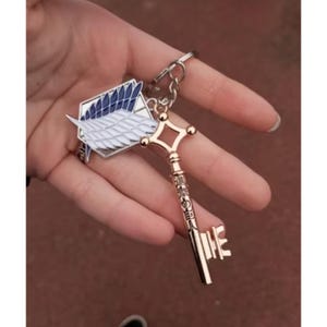 May include: A gold-colored key chain featuring a key and a badge with blue and white wings. The key has Japanese text and a decorative top. The badge has a white wing design with a blue background.