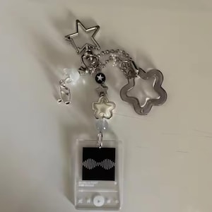 May include: A clear acrylic keychain with a music player design, featuring a black square with a sound wave graphic. The keychain includes silver star and flower charms, a musical note charm, and a chain with beads.