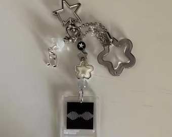 May include: A clear acrylic keychain with a music player design, featuring a black square with a sound wave graphic. The keychain includes silver star and flower charms, a musical note charm, and a chain with beads.