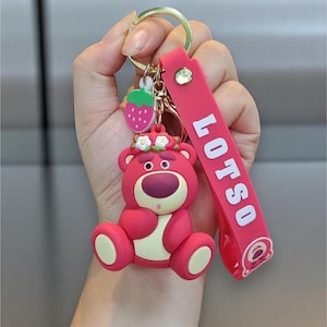 May include: A pink keychain featuring a cartoon bear character with a floral crown and a strawberry charm. The keychain has a pink strap with the word "LOTSO" in white. The bear has a cream-colored belly and feet.