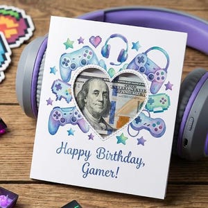 Könnte beinhalten: Eine Geburtstagskarte mit einem herzförmigen Ausschnitt, der einen 100-Dollar-Schein zeigt. Die Karte ist mit Gamecontrollern und dem Text "Happy Birthday, Gamer!" verziert. Ein lila-graues Headset liegt hinter der Karte, mit Pixel-Art-Aufklebern und Tastaturtasten verstreut.