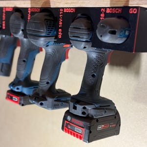 May include: A black wall-mounted tool storage rack holding five Bosch power drills. The rack features red printed text labels including Bosch, GSB 18V-110, and GO. The drills are blue and black with attached battery packs, hanging vertically from the rack.