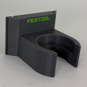Könnte beinhalten: Ein schwarzer, 3D-gedruckter Werkzeughalter mit dem grünen Schriftzug "FESTOOL". Der Halter hat eine kreisförmige Öffnung und eine rechteckige Basis, um ein Werkzeug zu halten. Der Halter besteht aus einem mattschwarzen Material.