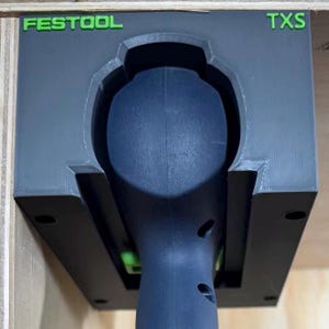 Puede incluir: Un soporte de herramientas gris y negro con la palabra "FESTOOL" en letras verdes en la parte superior izquierda y "TXS" en letras verdes en la parte superior derecha. Una herramienta negra está insertada en el soporte.
