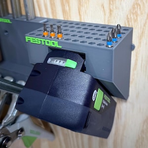 Puede incluir: Un organizador de herramientas Festool gris con una batería negra y varias brocas. El organizador tiene el nombre de la marca en letras verdes. La batería tiene un botón de encendido verde y un símbolo de Bluetooth.