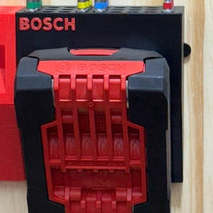 Könnte beinhalten: Schwarzes und rotes Bosch Elektrowerkzeugzubehör. Das Zubehör umfasst einen schwarzen Halter mit dem Markennamen "BOSCH" in Rot und einen roten Batteriehalter. Mehrere Bohrer sind in die Oberseite des Halters eingesetzt.
