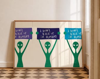 Op de afbeelding: Een ingelijste horizontale print met drie groene alienfiguren die elk een rechthoekig bord vasthouden. De borden zijn paars, donkerblauw en lichtblauw en tonen elk de witte tekst I DON'T BELIEVE IN HUMANS in een handgeschreven stijl.