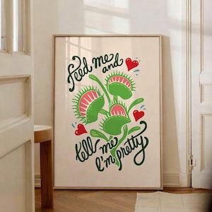 May include: A framed art print featuring a stylized Venus flytrap illustration. The artwork includes green flytraps with pink interiors and red hearts, accompanied by the text "Feed me and tell me I'm pretty" in a whimsical font.