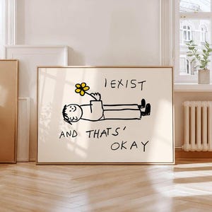May include: A rectangular wall art print featuring a black line drawing of a person lying down and holding a yellow flower. The text I EXIST AND THATS OKAY is written in black ink above and below the figure on a light cream background.