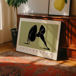 May include: A framed rectangular wall art print featuring a sage green background with a black abstract silhouette of a figure in a bent pose. The text S&middot;E&middot;L&middot;F&middot;B&middot;E&middot;L&middot;I&middot;E&middot;F is printed in black capital letters at the bottom of the print.