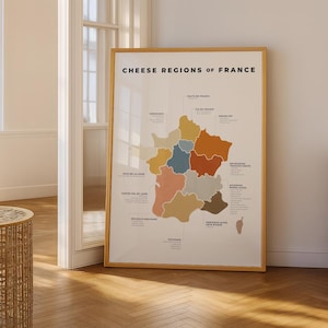 Puede incluir: Un póster enmarcado titulado "Cheese Regions of France" muestra un mapa colorido de Francia, destacando las regiones productoras de queso. El mapa presenta un fondo beige claro con regiones en tonos marrón, naranja, azul y rosa. El póster está en un marco de madera.