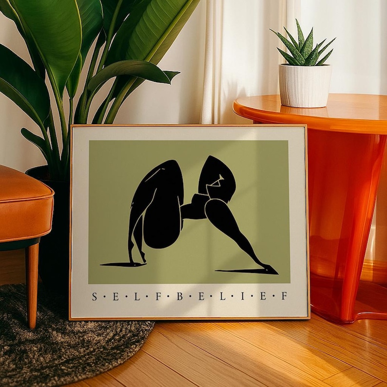 May include: A rectangular framed wall art print featuring a sage green background with a black abstract silhouette of a figure in a yoga pose. The text "S E L F B E L I E F" is printed in black capital letters at the bottom of the white border.