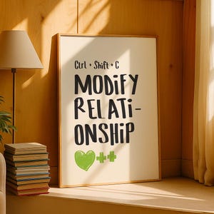 May include: A framed print with the words "Ctrl + Shift + C MODIFY RELATIONSHIP" in black, with a green heart and two green plus signs. The print is in a wooden frame and is leaning against a wall. A stack of books is visible in the foreground.