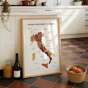 Pode incluir: Uma impressão emoldurada intitulada "PIZZA REGIONS OF ITALY" mostra um mapa da Itália, com regiões codificadas por cores. A impressão está em uma moldura de madeira clara, encostada em um armário branco. Uma cesta de cebolas, uma garrafa de vinho e um frasco estão em primeiro plano.