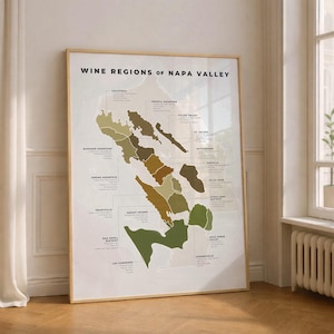 May include: A framed poster titled "Wine Regions of Napa Valley" displays a map of the region, highlighting various sub-appellations with different colors. The poster is in a light-colored wooden frame and leans against a white wall.
