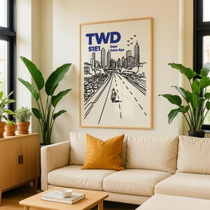 May include: A framed art print featuring a black and white illustration of a city skyline and a road with a cyclist. The print has the text "TWD S1E1 Days Gone Bye" in blue. The artwork is displayed above a beige sofa with an orange pillow.