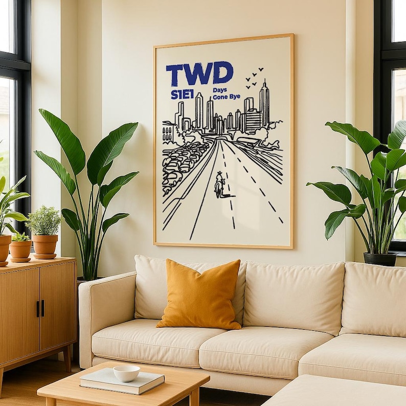 May include: A framed art print featuring a black and white illustration of a city skyline and a road with a cyclist. The print has the text "TWD S1E1 Days Gone Bye" in blue. The artwork is displayed above a beige sofa with an orange pillow.