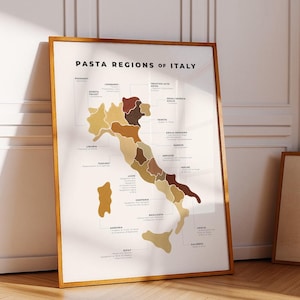 May include: A framed print titled "PASTA REGIONS OF ITALY" displays a map of Italy, segmented into regions with varying shades of brown and beige. Text labels identify regions such as Piedmont, Lombardy, and Tuscany. The frame is a light brown.
