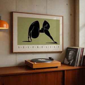 May include: A framed rectangular wall art print featuring a stylized black silhouette of a person in a yoga pose against a muted olive green background. The text S E L F B E L I E F is printed in black capital letters centered below the illustration.