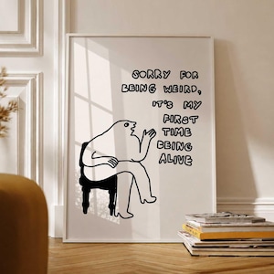May include: A framed art print with a minimalist black and white illustration. The artwork features a seated figure with the text "Sorry for being weird, it's my first time being alive." The print is displayed on a white wall.
