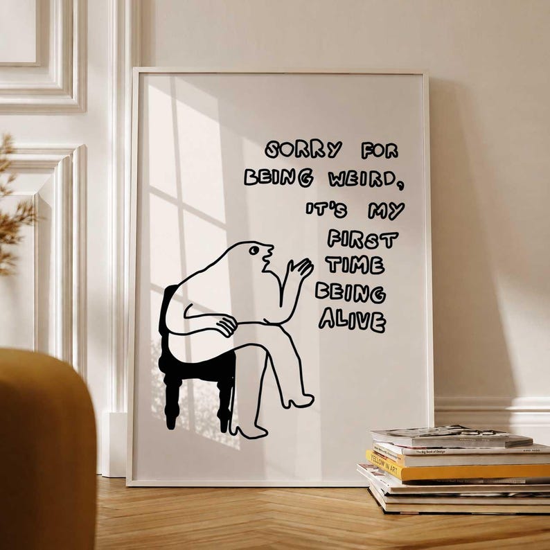 May include: A framed art print with a minimalist black and white illustration. The artwork features a seated figure with the text "Sorry for being weird, it's my first time being alive." The print is displayed on a white wall.