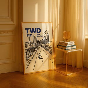 May include: A framed art print with the text "TWD S1E1 Days Gone Bye" in blue. The artwork depicts a road leading to a city skyline, with a figure on a motorcycle. A glass side table holds books and a decorative vase.